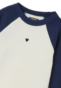 Weißes, geripptes Langarmshirt mit marineblauen Raglanärmeln und einer kleinen marineblauen Herzstickerei auf der Brust. OVS-Markenetikett ist innen sichtbar.