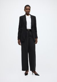 Μαύρο ραμμένο σακάκι πάνω από λευκό πουκάμισο με κουμπιά, συνδυασμένο με μαύρο παντελόνι με φαρδιά μπατζάκια και μαύρα τακούνια μυτερά στο άκρο. Λείες υφές σε όλο το σύνολο.