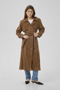 Jonge vrouw die een riem vastmaakt aan een lange bruine suède trenchcoat, draagt blauwe jeans en zwarte schoenen met een laag hakje en een spitse neus, op een grijze achtergrond.