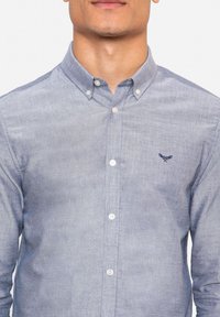 Lange mouwen knoopshirt van lichtblauwe chambray stof. Bevat een kraag, knopen aan de voorkant en een klein geborduurd logo op de borst.