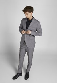 Costume gris clair avec un col cranté, à simple boutonnage, deux poches avant, accompagné d'une chemise foncée et de bottines noires.