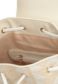 Sac seau en tissu avec un extérieur tissé couleur beige, des accents blancs et une quincaillerie dorée. Comprend un intérieur beige lisse avec une poche zippée.
