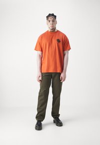 Camiseta de algodón naranja con un gráfico negro en el pecho, combinada con pantalones verde oliva y zapatos negros con cordones.
