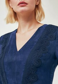 Mörkblå klänning med V-ringning, med intrikata spetsdetaljer längs sidan. Mjuk, mockaliknande textur med slät yta.