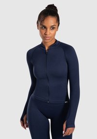 Chaqueta atlética azul marino con mangas largas, diseño ajustado, cierre de cremallera y textura acanalada. Usada con leggings a juego para un look deportivo.