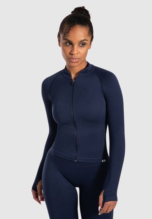 Marineblaues Sportjackett mit langen Ärmeln, tailliertem Schnitt, Reißverschluss und geribbelter Textur. Wird mit passender Leggings für einen sportlichen Look getragen.