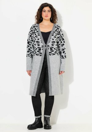 Grauer gestrickter Cardigan mit schwarzem Leopardenmuster, Frontknöpfen und gerippten Bündchen, kombiniert mit schwarzen Jeans und klobigen schwarzen Stiefeln.