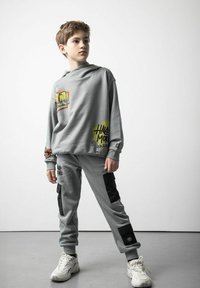 Szary zestaw bluzy z kapturem i spodni dresowych z graficznymi nadrukami, czarnymi akcentami i luźnym krojem. Wykonany z miękkiego materiału. Białe sneakersy.