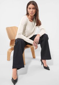 Frau sitzt auf einem Holzstuhl, trägt einen cremefarbenen Pullover, schwarze weitgeschnittene Hosen und schwarze spitze High Heels mit Schnallen.