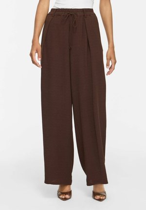Frau trägt hoch taillierte, locker sitzende braune Hose mit Kordelzug in der Taille und Falten, kombiniert mit braunen offenen Absätzen und weißem Oberteil.
