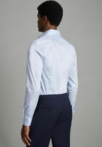 Chemise bleu clair à manches longues, avec un col évasé et des boutons blancs, associée à un pantalon bleu marine, vue de dos.