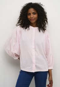 Light pink, semi-sheer blouse with puffed sleeves and dotted texture. Features a button-down front and rounded hem. Paired with blue jeans.
