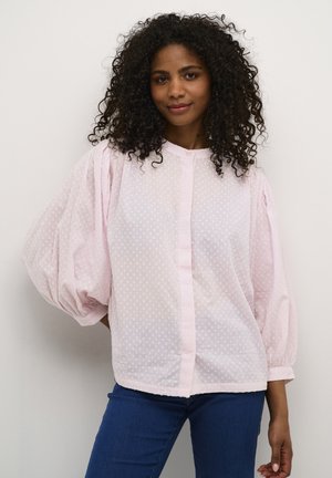 Blouse rose clair semi-transparente avec des manches bouffantes et une texture à pois. Elle présente un boutonnage sur le devant et un ourlet arrondi. Associée à un jean bleu.