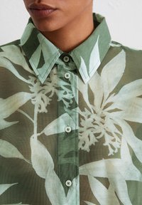 Chemise à motif floral vert avec un devant boutonné, un col et un tissu léger. Présente des feuilles translucides et des boutons dans une teinte assortie.