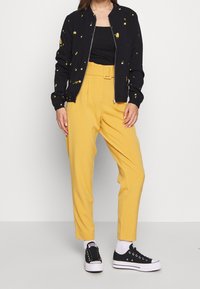 Černá bomber bunda se žlutými květinovými vzory, zipový uzávěr, v kombinaci s vysokými žlutými kalhotami a černými teniskami. Bílé ponožky jsou viditelné.