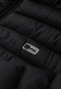 Tejido de chaqueta acolchada negra con textura de acolchado, que presenta un pequeño parche de logo plateado. El material parece suave y resistente al agua.