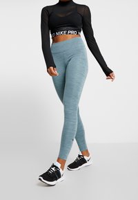 Černý dlouhý crop top s logem na pásku, v kombinaci se světle modrými mramorovanými legínami. Modelka má na nohou černé sportovní boty a stojí v profilu.