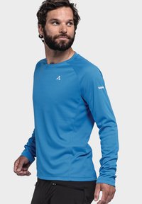 Blaues langärmliges Sporthemd aus strukturiertem Stoff, mit Rundhalsausschnitt und einem kleinen Logo auf der Schulter. Getragen mit schwarzen Hosen.