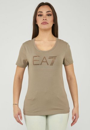Vrouw met lang bruin haar die een beige T-shirt draagt met de letters "EA7" in kleine bruine studs op de borst.