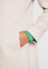 Mano nella tasca di un cappotto beige con la manica di una camicia verde visibile al polso, evidenziando la texture del tessuto e il contrasto di colori.