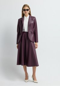 Ensemble en cuir violet composé d'un blazer et d'une jupe avec une texture lisse, associé à une chemise blanche et des lunettes de soleil noires. Chaussures à talons hauts blanches.