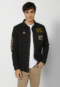Camisa negra de botones con mangas largas, que presenta texto amarillo, un parche con logo y un tejido texturizado. Llevada sobre una camiseta blanca.