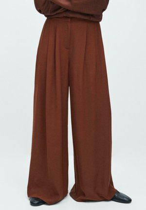 Pantalon marron à jambes larges porté avec des chaussures noires pointues, assorti à un haut marron à manches longues, bras croisés.