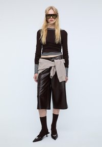 Schwarzes Langarmshirt mit weißen Streifen, braune Leder-Culottes, beiger Pullover um die Taille gebunden und spitze dunkle Kitten-Heels. Grüne Sonnenbrille.