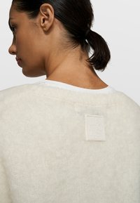 Mujer con cabello oscuro recogido que lleva un suéter blanco suave y borroso con un parche cuadrado blanco en la parte superior de la espalda.