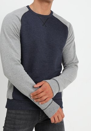 Mann in einem navyblauen und hellgrauen Langarm-Sweatshirt, der mit verschränkten Armen die Ärmel anpasst, vor einem schlichten weißen Hintergrund.