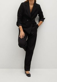 Žena v černé dvouřadé saku s vyhrnutými rukávy, černých kalhotách a černých balerínách, stojící na pozadí světlé holé stěny.