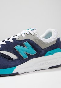 Siniset mokkakengät harmaan ja turkoosin yksityiskohtien kanssa, valkoinen verkotettu kieli, paksu valkoinen välipohja, ja 'new balance' -brändäys.