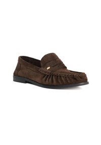 Bruine suede instappers met een rafelige rand, decoratieve stiksels en een klein gouden embleem aan de zijkant; ronde neus en platte rubberen zool.