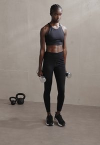 Juoda sportinė trumpa palaidinė ir aukštu juosmeniu leggingsai, laikantys sidabro spalvos hantelius, juodi sportbačiai, su girietėmis fone ant šviesaus grindų.