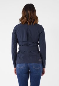Giacca impermeabile blu navy con vita con cintura, dettaglio con piega sul retro e maniche lunghe; visibile da dietro su uno sfondo bianco semplice.