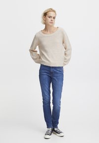 Helle beige Strickjacke mit weitem Ausschnitt, kombiniert mit schmal geschnittenen blauen Jeans und schwarzen Leinenschuhen. Einfaches, strukturiertes Design.