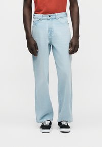 Ανοιχτό μπλε τζιν παντελόνι με φαρδιά μπατζάκια και σχέδιο με πέντε τσέπες, με εμφανείς ραφές· συνδυασμένο με μαύρα αθλητικά παπούτσια με λευκά κορδόνια.