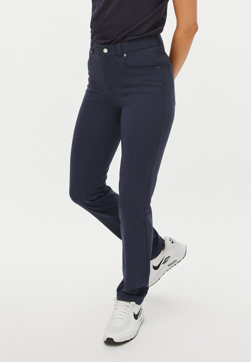 Navyblauwe, slim-fit broek van rekbare stof. Met een klassieke vijf-pocket ontwerp en een gladde textuur. Draagt met witte sneakers.