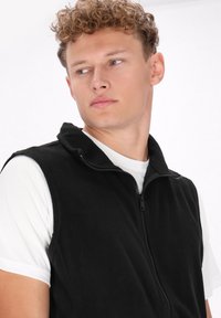 Schwarze Fleece-Weste mit Reißverschluss vorne, Stehkragen und ohne Ärmel, getragen über einem weißen T-Shirt. Glatte Textur und lässiges Design.