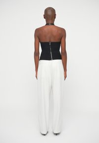 Pessoa com cabeça rapada, vestindo um top preto sem alças com fecho atrás e calças largas listradas em branco, de pé contra um fundo branco.