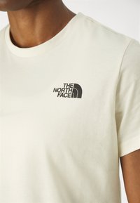 Detailní záběr na osobu oblečenou v krémovém tričku s černým logem "The North Face" na levé horní části hrudi.