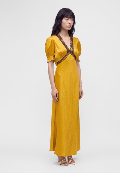 Femme portant une longue robe jaune moutarde à manches bouffantes et bordure en dentelle noire, debout les bras détendus le long du corps devant un fond uni.