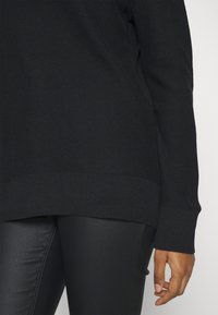 Pull noir texturé avec un ourlet et des poignets côtelés, porté avec un pantalon noir ajusté. Accent sur le motif tricoté et la finition du matériau.
