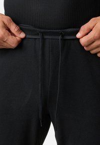 Zwarte fleece joggers met een gestructureerde afwerking, voorzien van een flexibele tailleband en verstelbaar koord. Handen grijpen de tailleband.