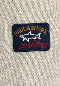 Tessuto lavorato a maglia beige con una toppa in tessuto blu navy. La toppa presenta "PAUL & SHARK" in oro e "yachting" in rosso, con due sagome di squalo bianche.
