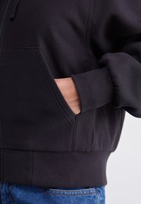 Hoodie zippé noir en tissu doux, doté d'une poche avant, de poignets et d'un ourlet côtelés. La vue latérale met en avant la poche et les textures.