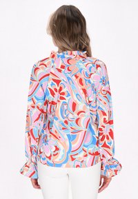 Bluză lungă, colorată, cu mânecă lungă, având un model floral în nuanțe de roz, roșu și albastru. Prezintă un guler volanat și manșete adunate. Textură de țesătură netedă.