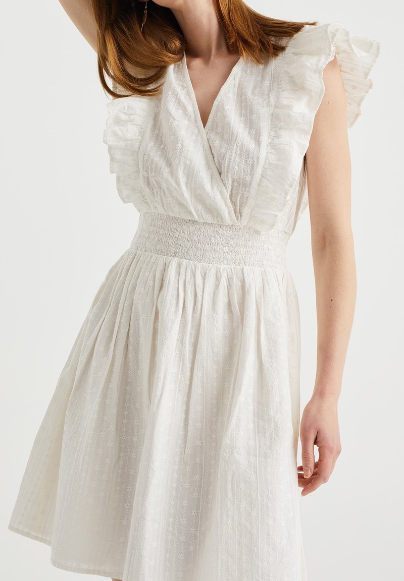 Robe blanche avec des manches courtes à volants, un décolleté en V, une taille smockée et une jupe évasée, présentant un tissu texturé avec des motifs subtils.