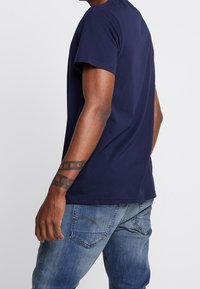 En man klädd i en marinblå kortärmad skjorta och blå jeans, som visar en geometrisk svart tatuering på sin vänstra underarm.