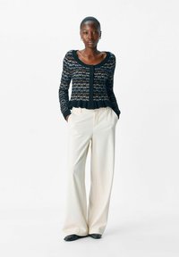 Cardigan noir à motifs avec un ourlet volanté, associé à un pantalon beige à jambes larges et des chaussures noires. Le cardigan présente un devant à boutons.